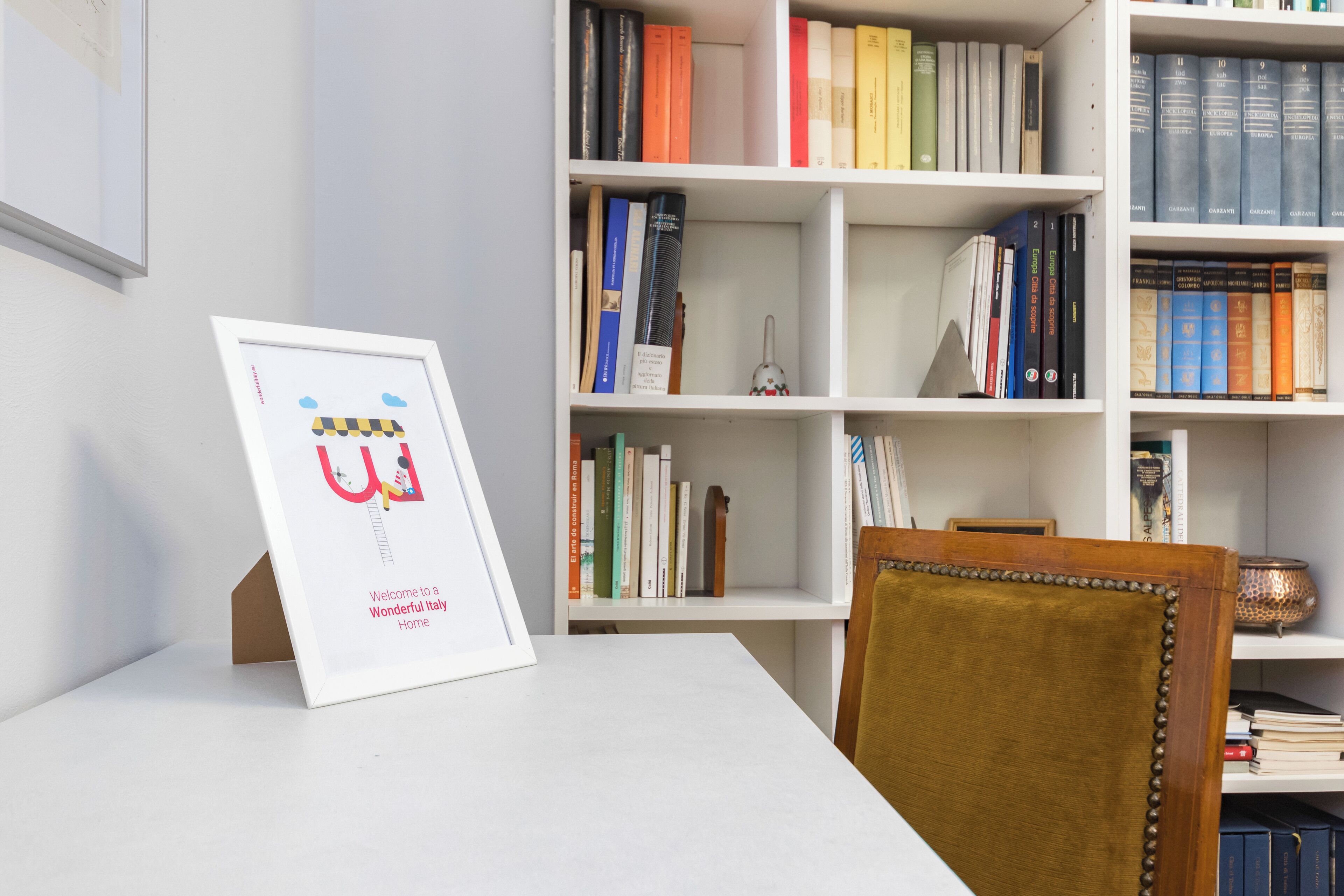Ein gerahmtes Willkommen-Schild, ein Bücherregal mit unterschiedlichen Büchern und ein hölzerner Stuhl.