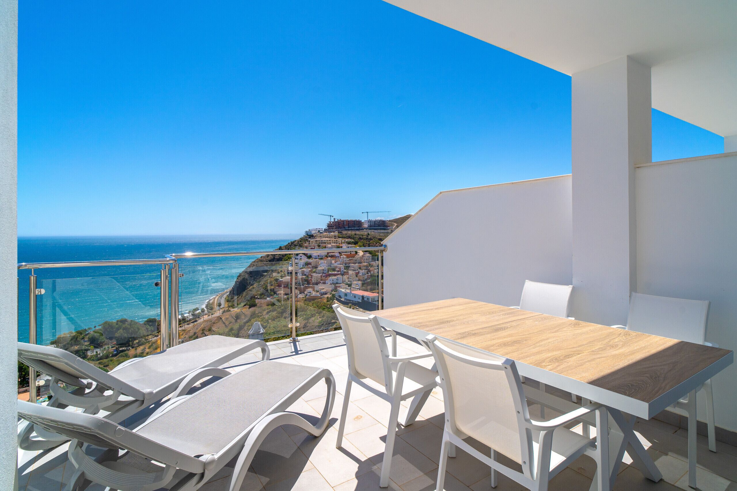 Un balcone con un tavolo di legno e sedie bianche, vista su una cittadina costiera e il mare.