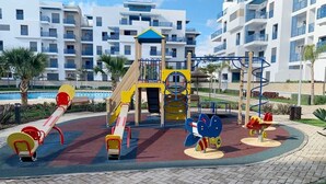 Zona de jocs amb tobogans i estructures d'escalada, envoltada d'una piscina i palmeres, amb un edifici modern al fons.