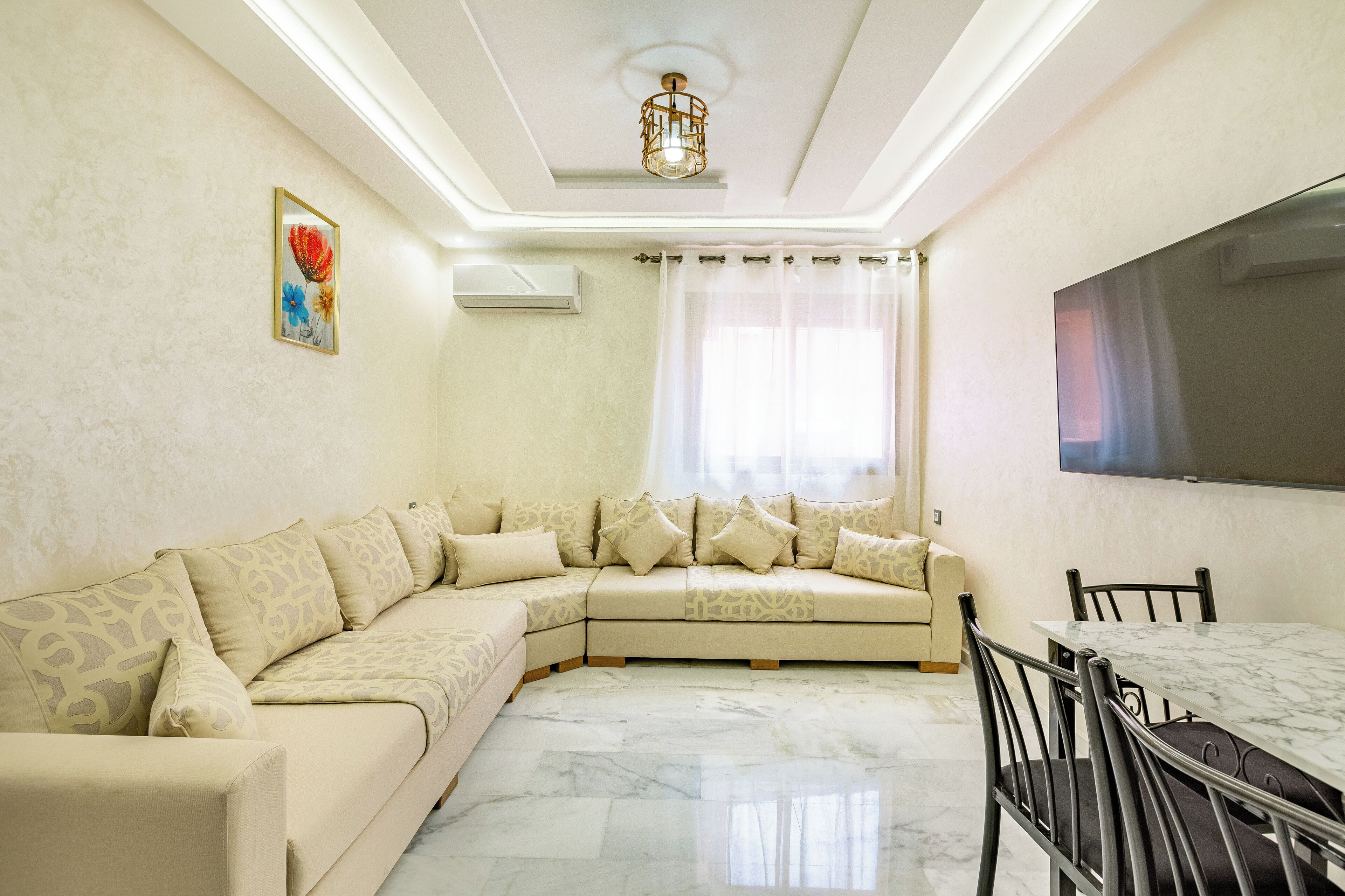 A living room with a beige sectional sofa, a marble floor, a dining table with chairs, a flat-screen TV mounted on the wall, and a window with sheer curtains.