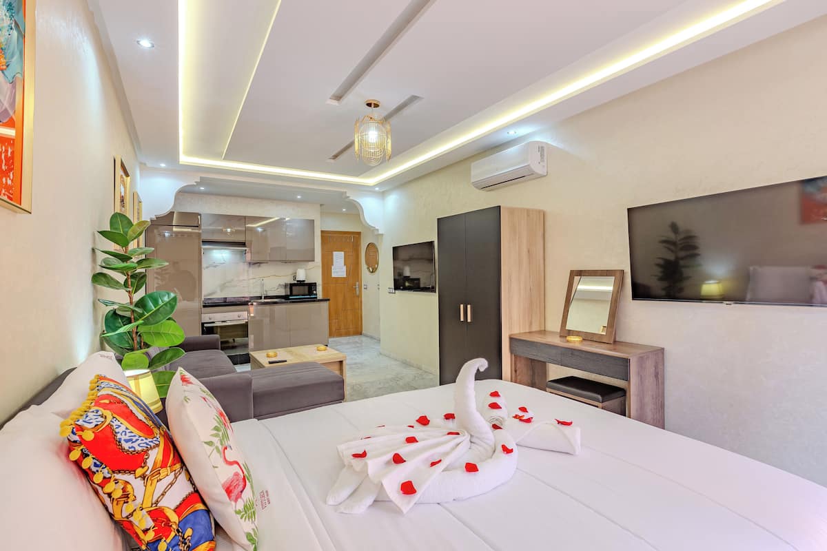 A modern hotel room with a neatly made bed, a dining area in the background, and a TV mounted on the wall.
