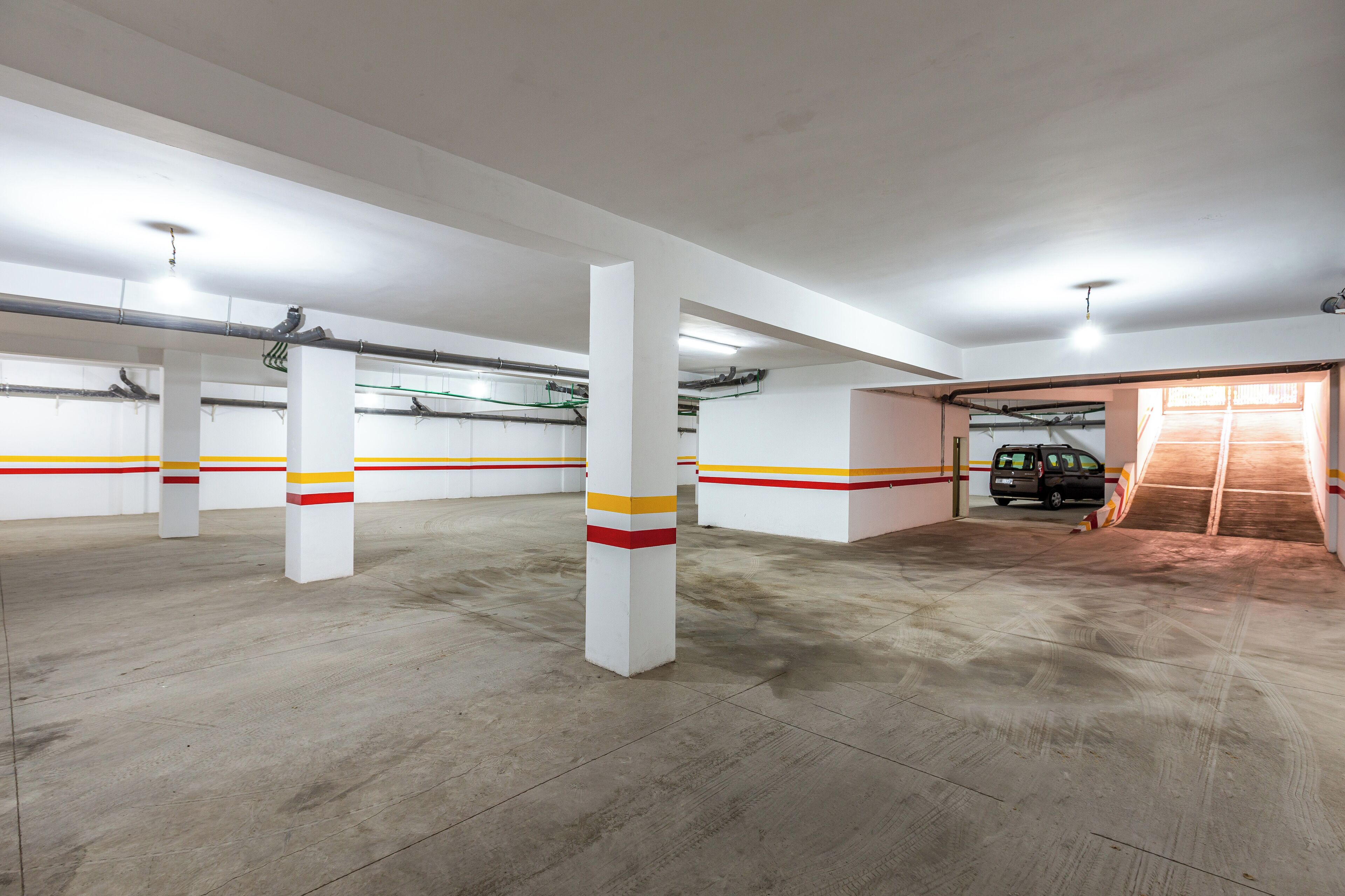 A spacious parking garage with white pillars, red and yellow stripes, and a car parked in the distance.