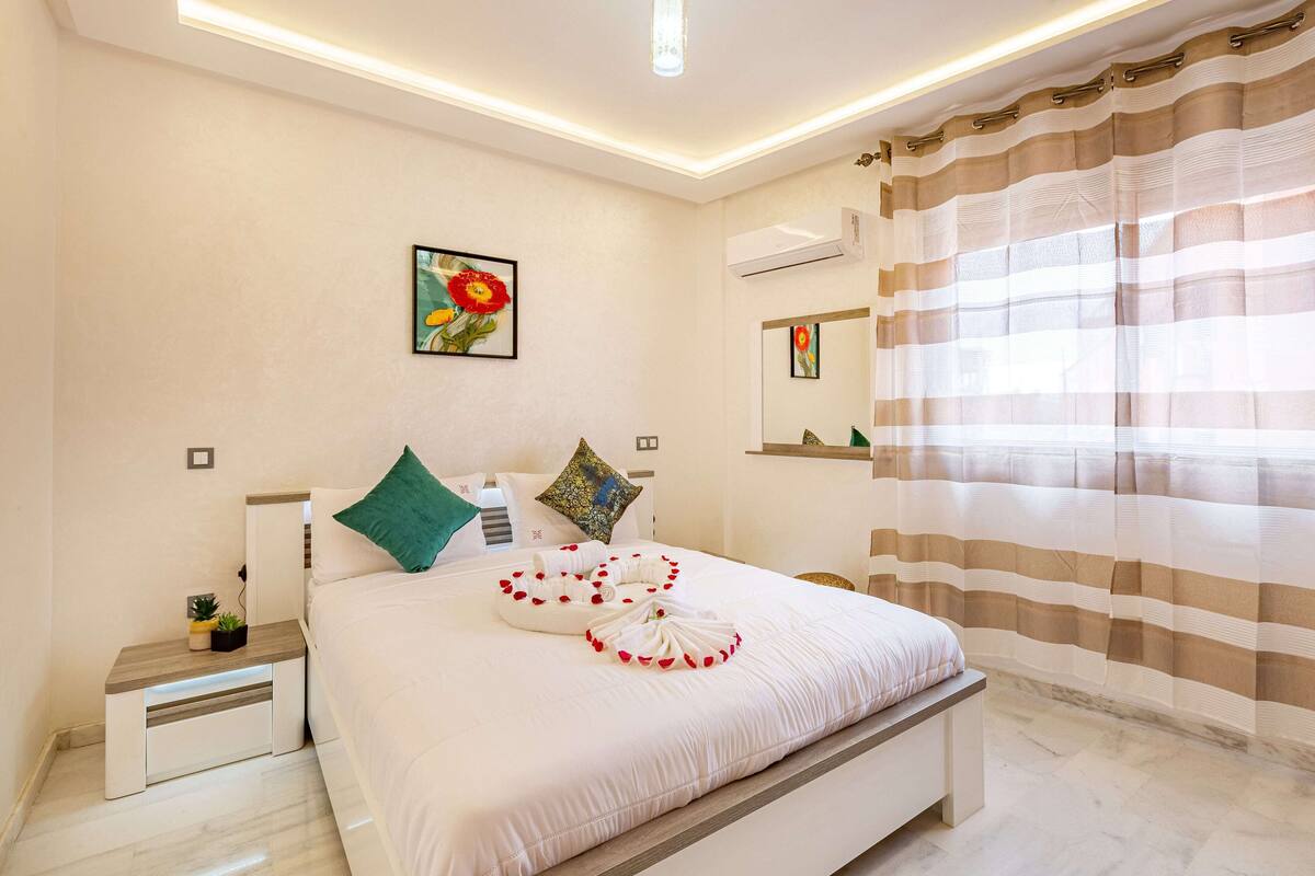 A neatly made bed with a pillow arrangement, a bedside table with a plant, and a painting on the wall.