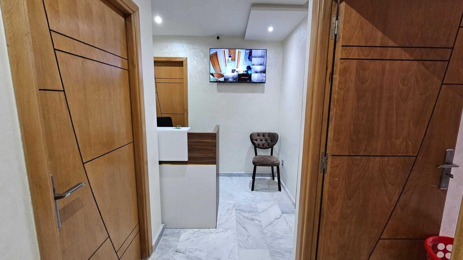 Un couloir avec des portes en bois, un petit coin salon, un téléviseur fixé au mur et un sol carrelé.