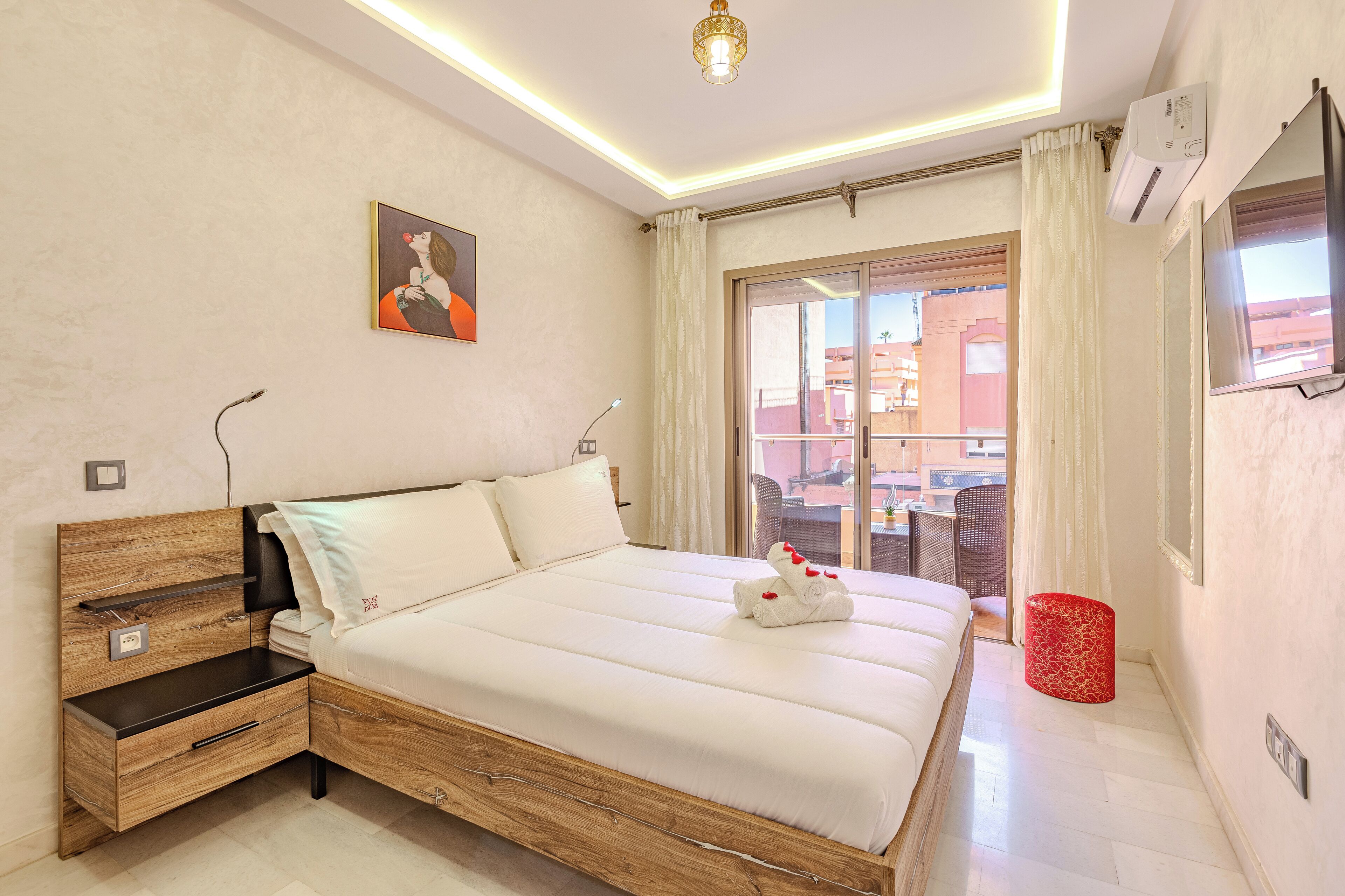 A bedroom with a wooden bed frame, white bedding, a bedside table with a lamp, a red trash bin, and a window with curtains.