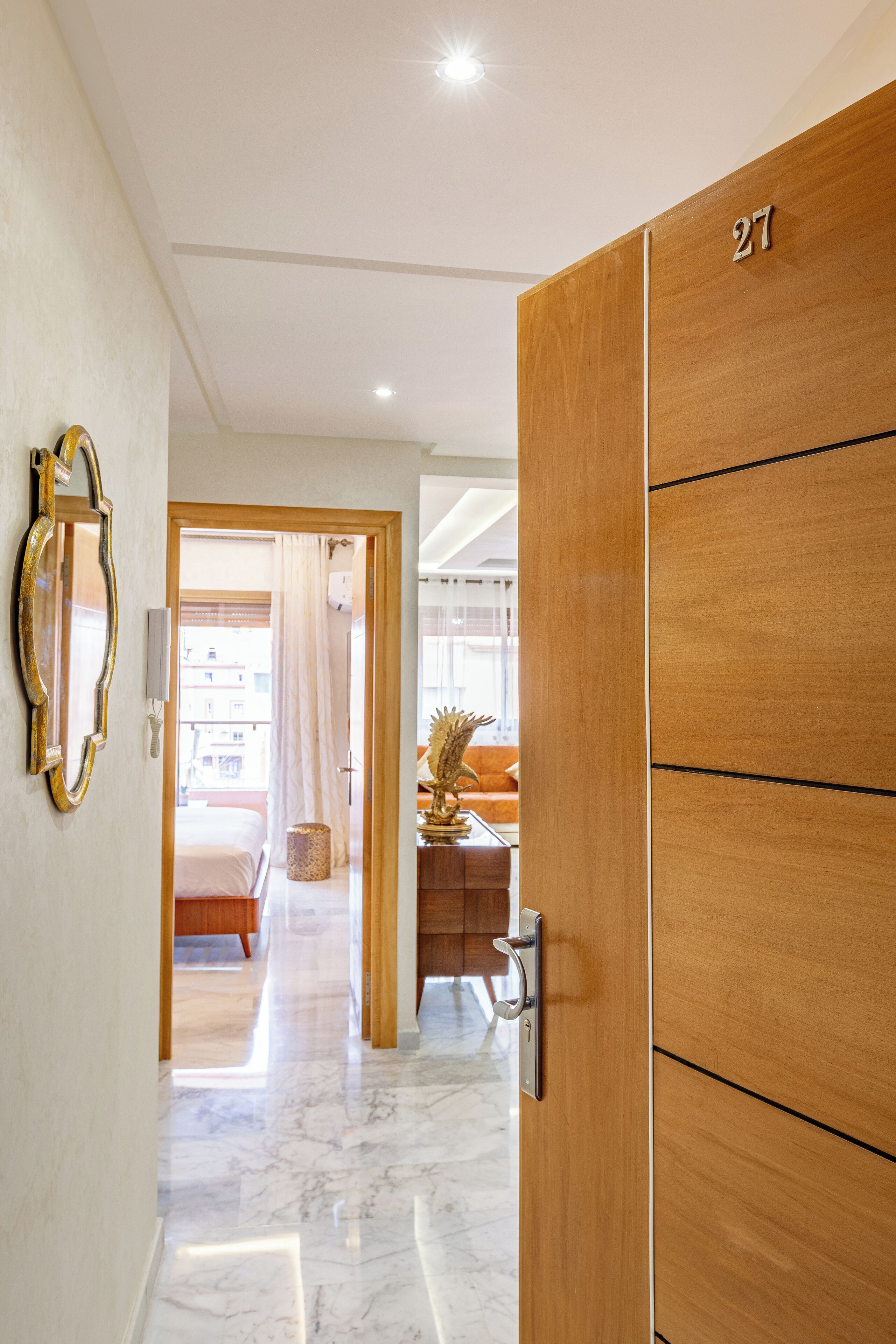 Habitación de hotel con puerta de madera marcada con el número "27", suelo de mármol, una cama, un escritorio con una planta y vistas al balcón exterior.