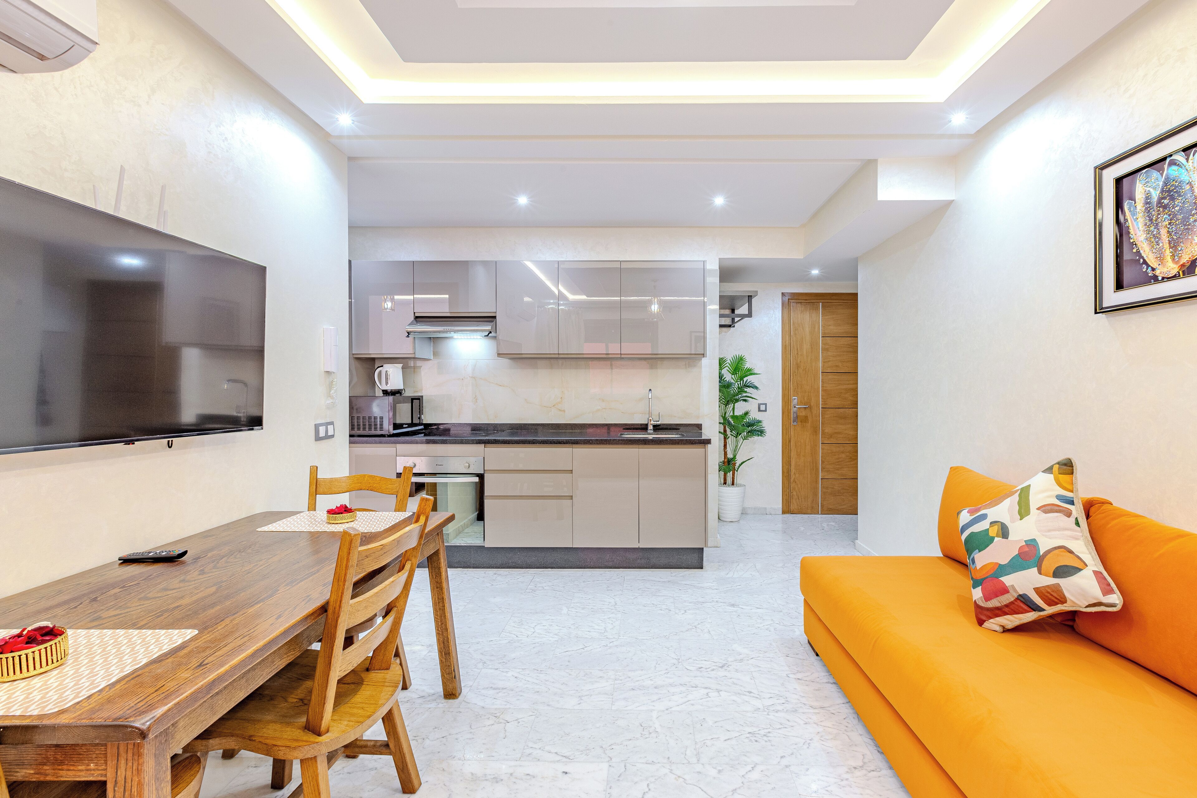 Een moderne woonkamer met een houten eettafel, een oranje bank en een flatscreen televisie die aan de muur is bevestigd.