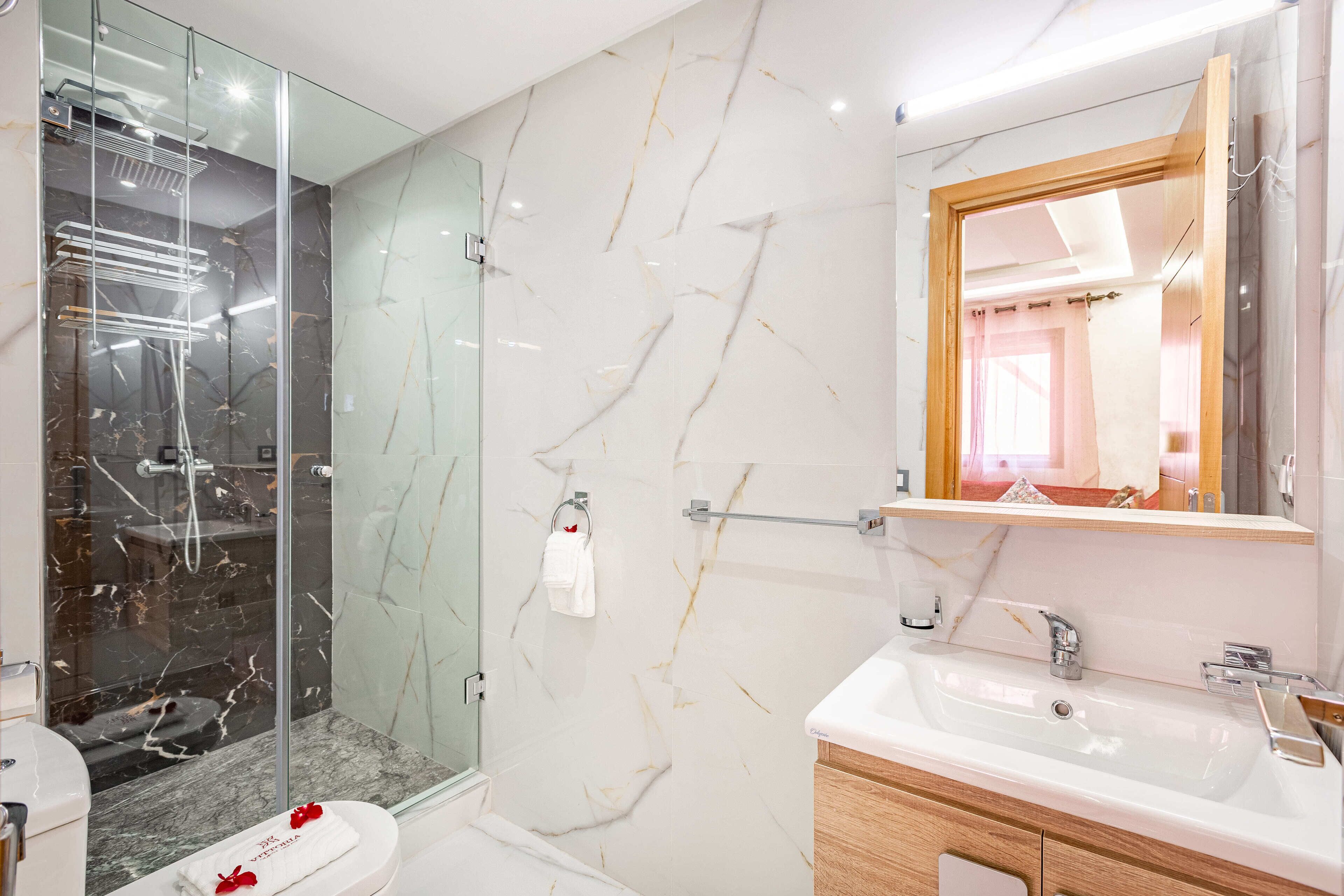 A modern bathroom with a glass-enclosed shower, marble walls, a wooden vanity, and a large mirror.