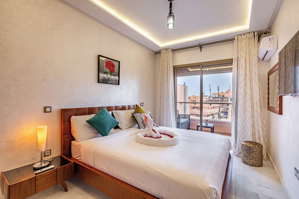 A hotel room with a large bed, a bedside table with a lamp, a wall-mounted air conditioner, a window with curtains, and a city view.