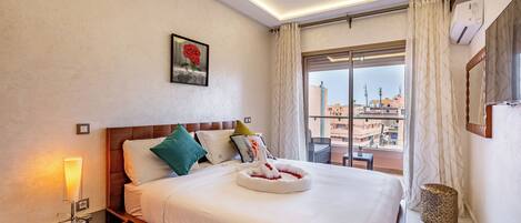 A hotel room with a large bed, a bedside table with a lamp, a wall-mounted air conditioner, a window with curtains, and a city view.