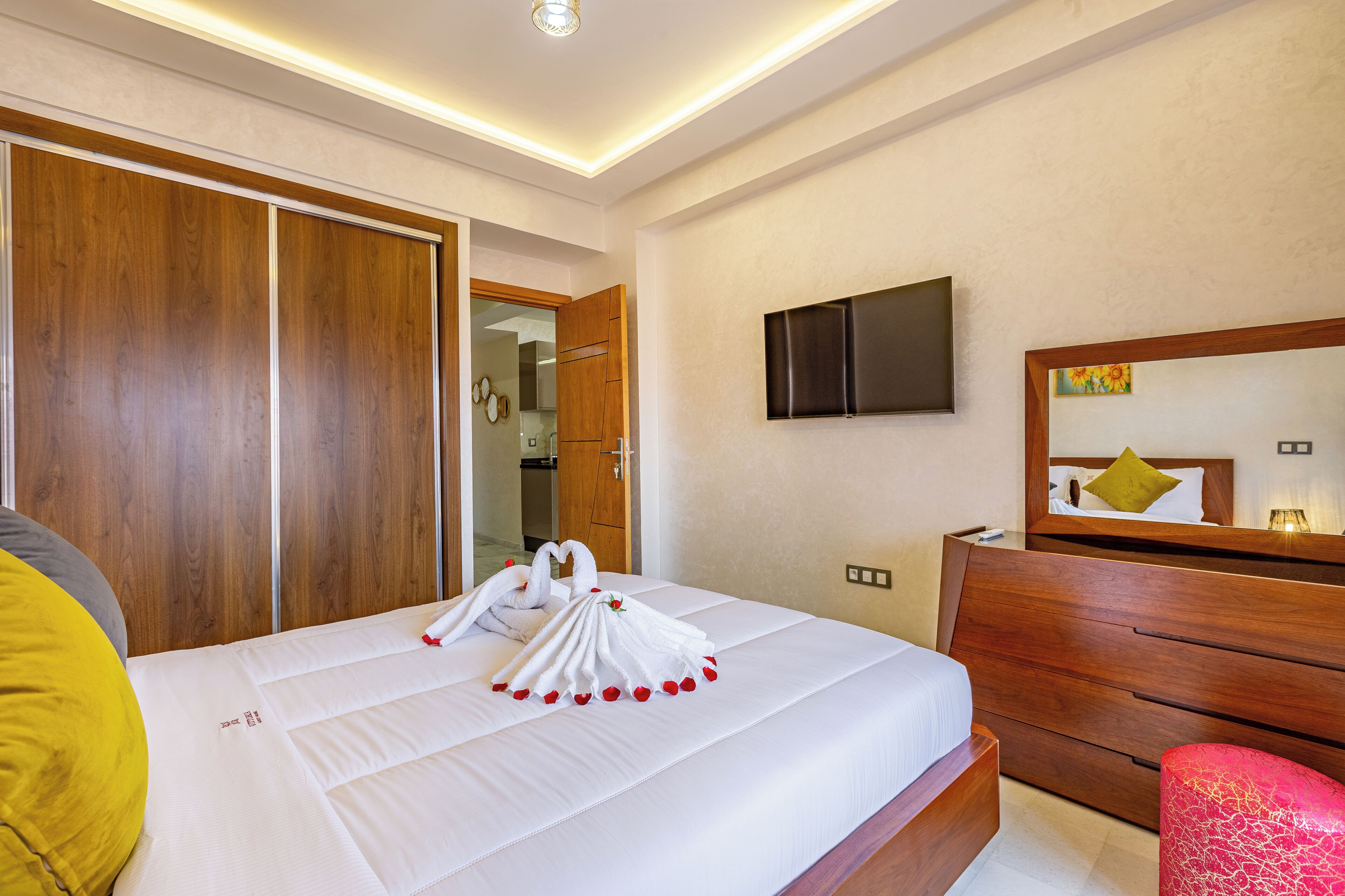 A hotel room with a neatly made bed, a wooden wardrobe, a television mounted on the wall, and a mirror above the dresser.
