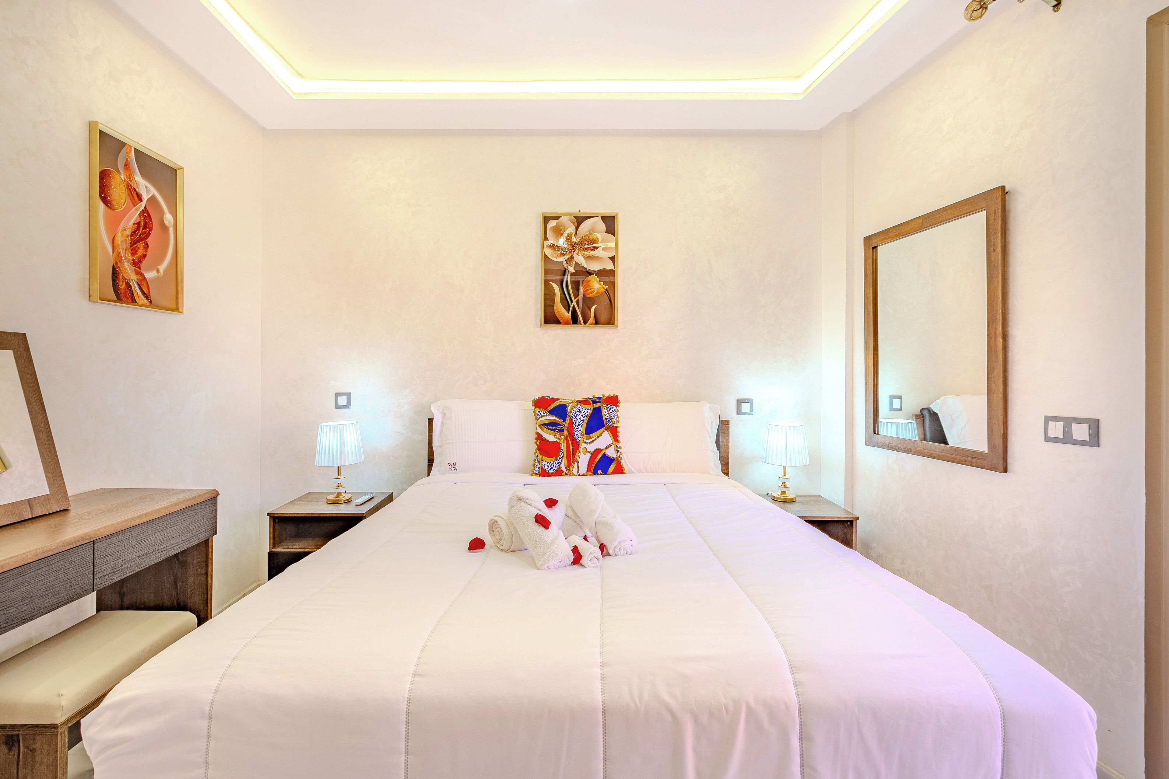 Quarto de hotel com uma cama grande, duas mesas de cabeceira com luminárias, um banco e obras de arte nas paredes.