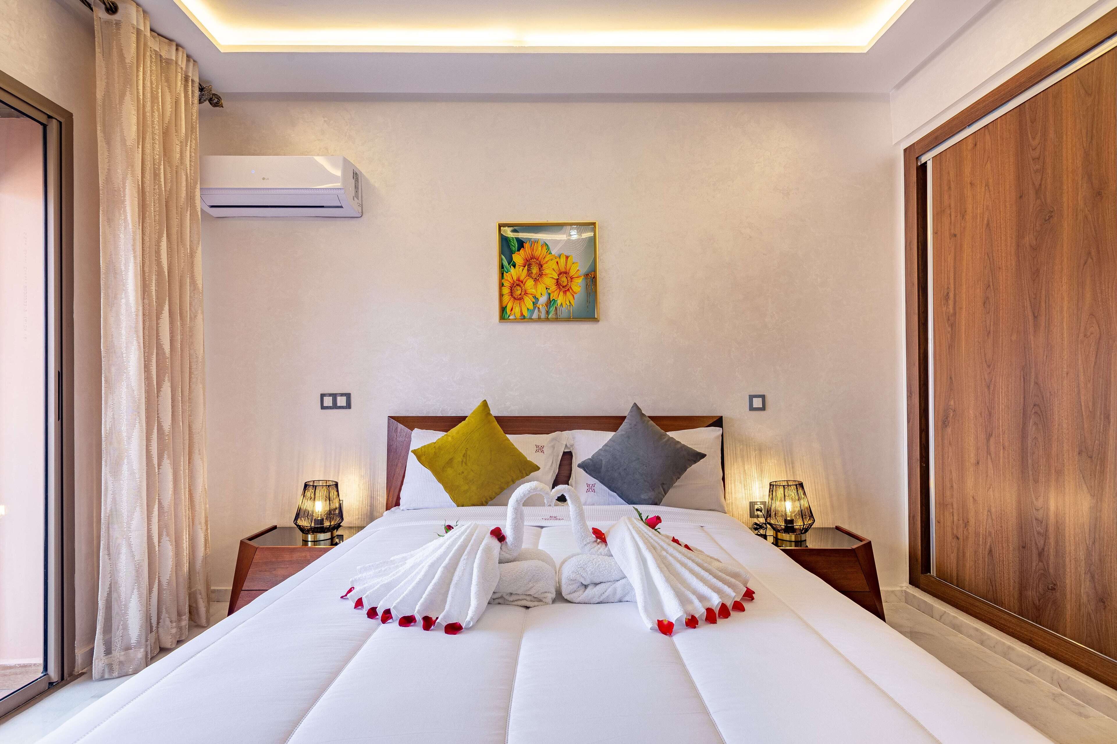 A neatly made bed with towels arranged in swan shapes, a painting of sunflowers on the wall, and a wooden panel on the right.