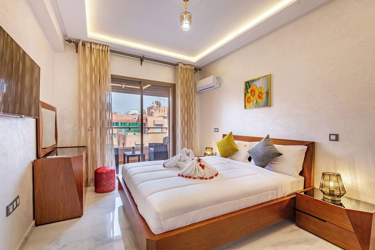 A hotel room with a large bed, a wooden desk, a red trash bin, and a view of buildings outside.