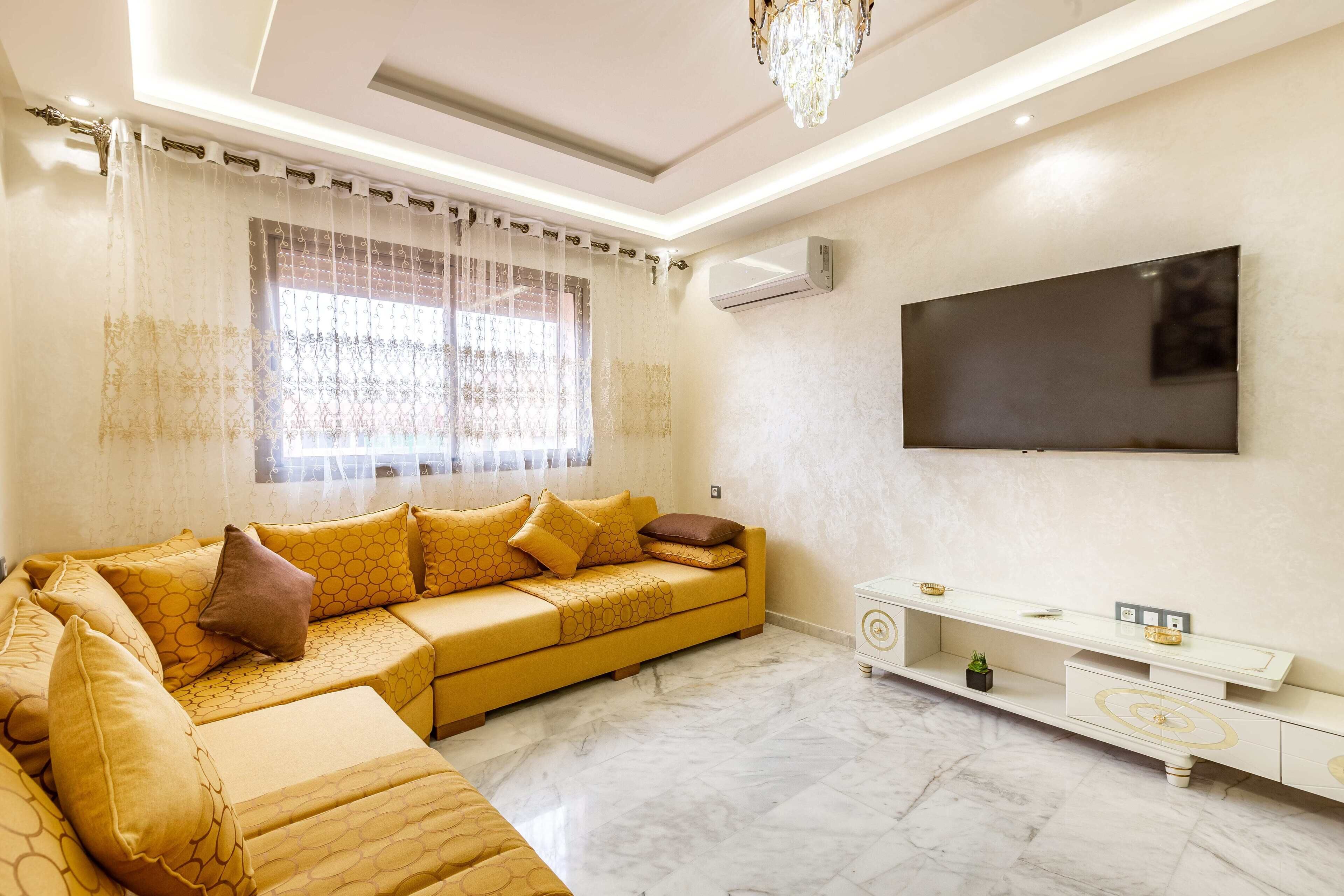 A modern living room with a yellow sectional sofa, a flat-screen TV mounted on the wall, and a white console table with a small plant.