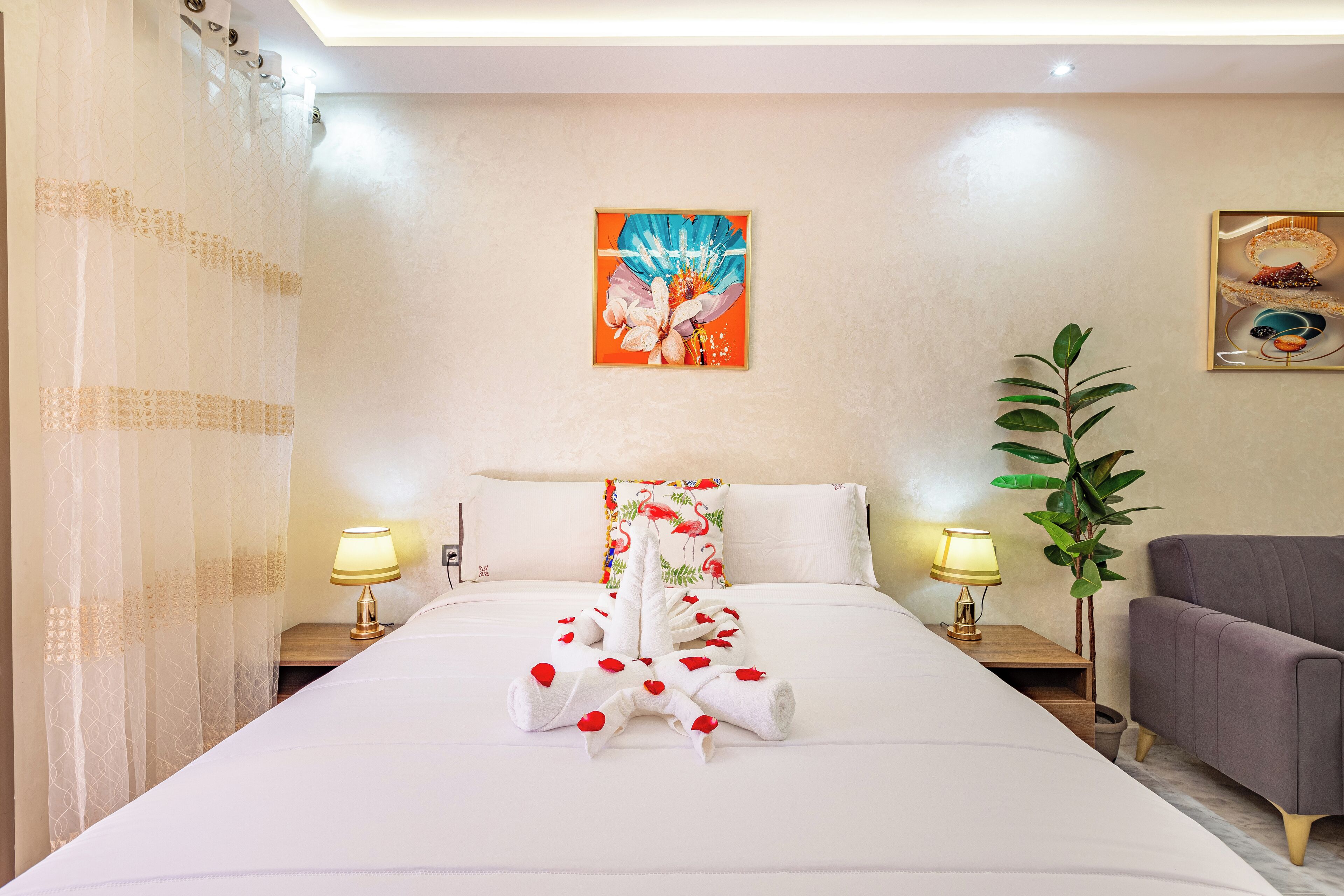 A hotel room with a neatly made bed, two bedside lamps, a plant, and artwork on the walls.