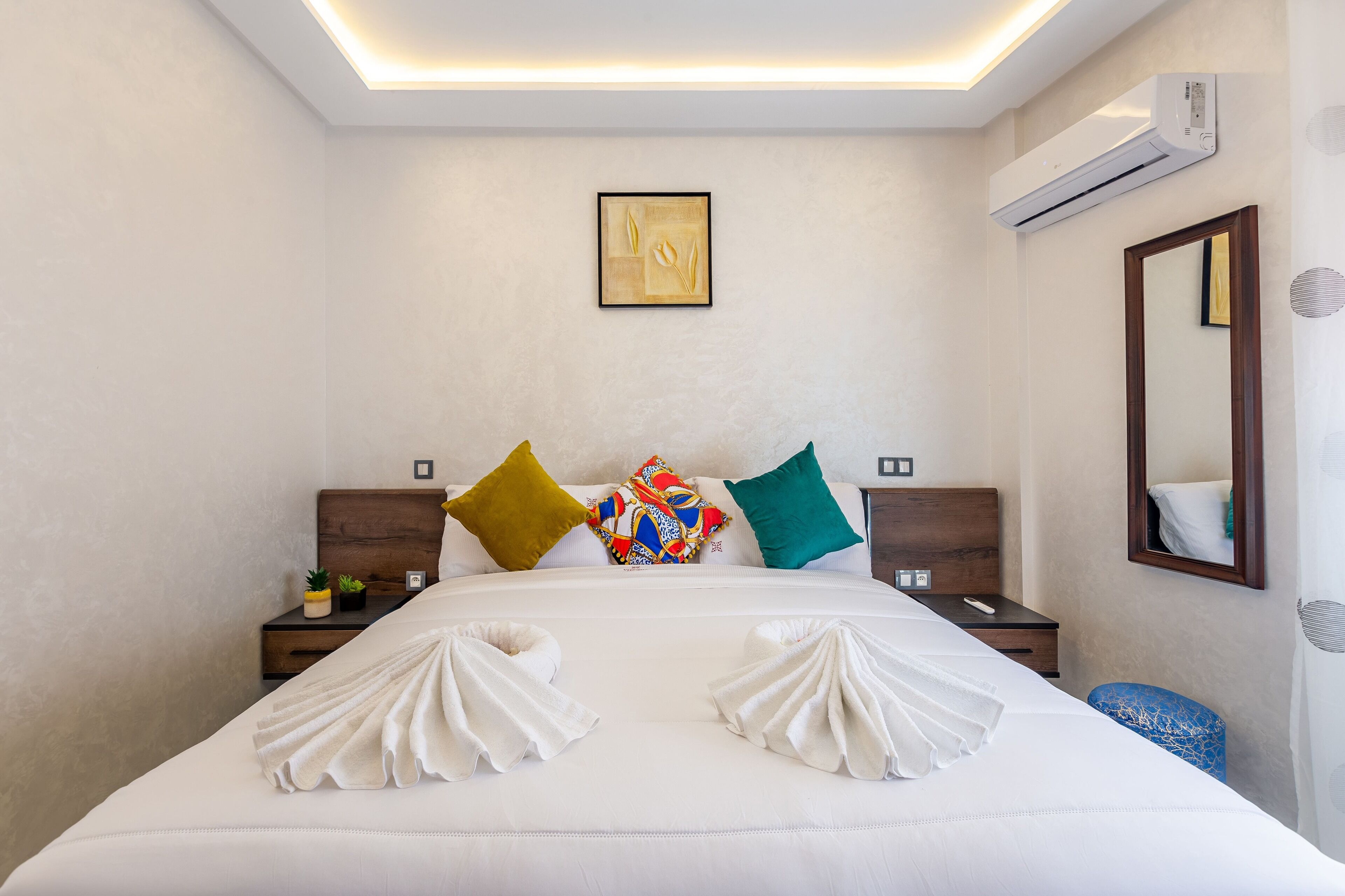 A hotel room with a neatly made bed, a bedside table with a lamp, a wall-mounted air conditioner, and a mirror.