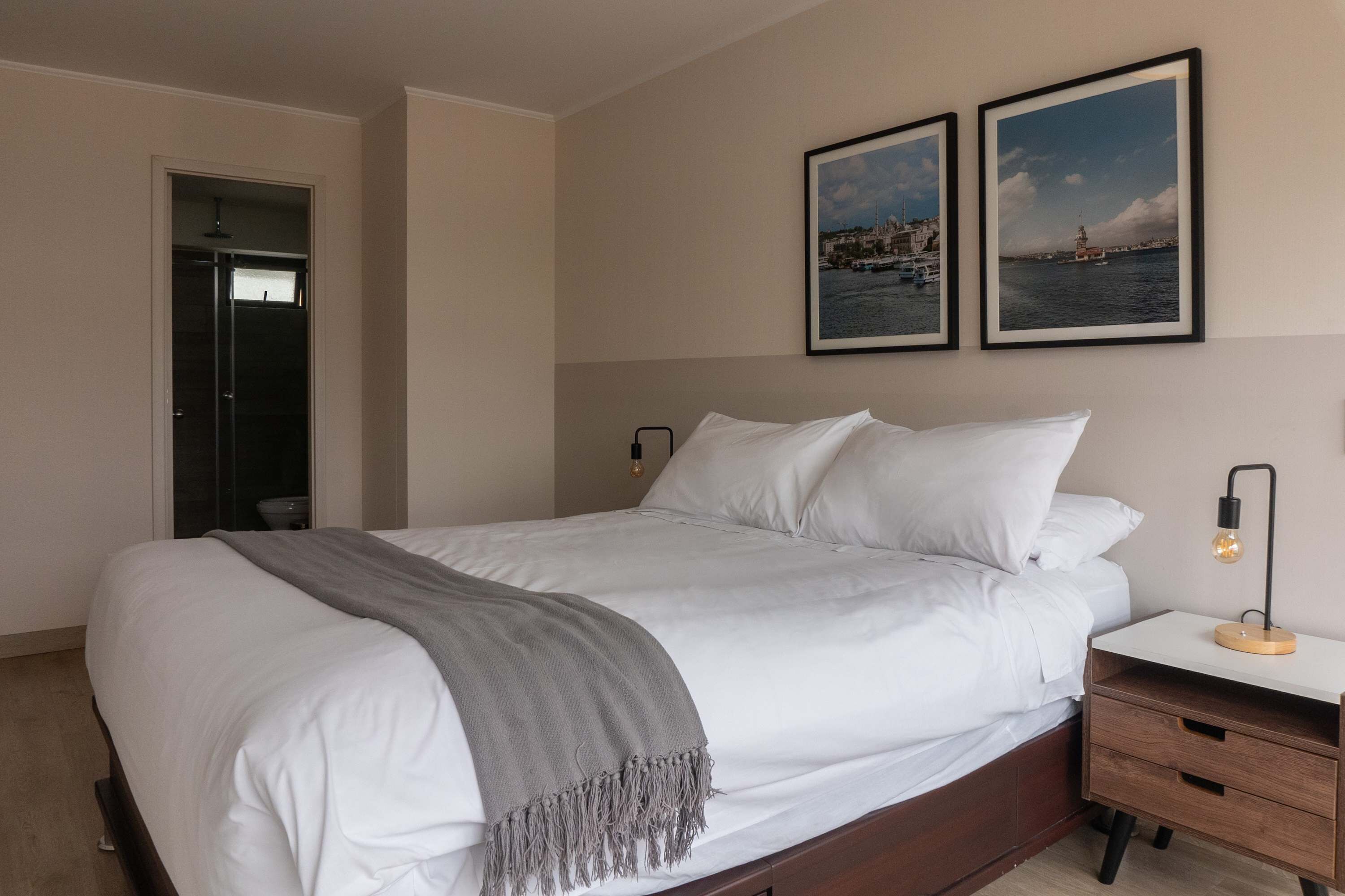 Un lit bien fait, recouvert d’un plaid gris, une table de chevet avec une lampe, et deux tableaux encadrés accrochés au mur.