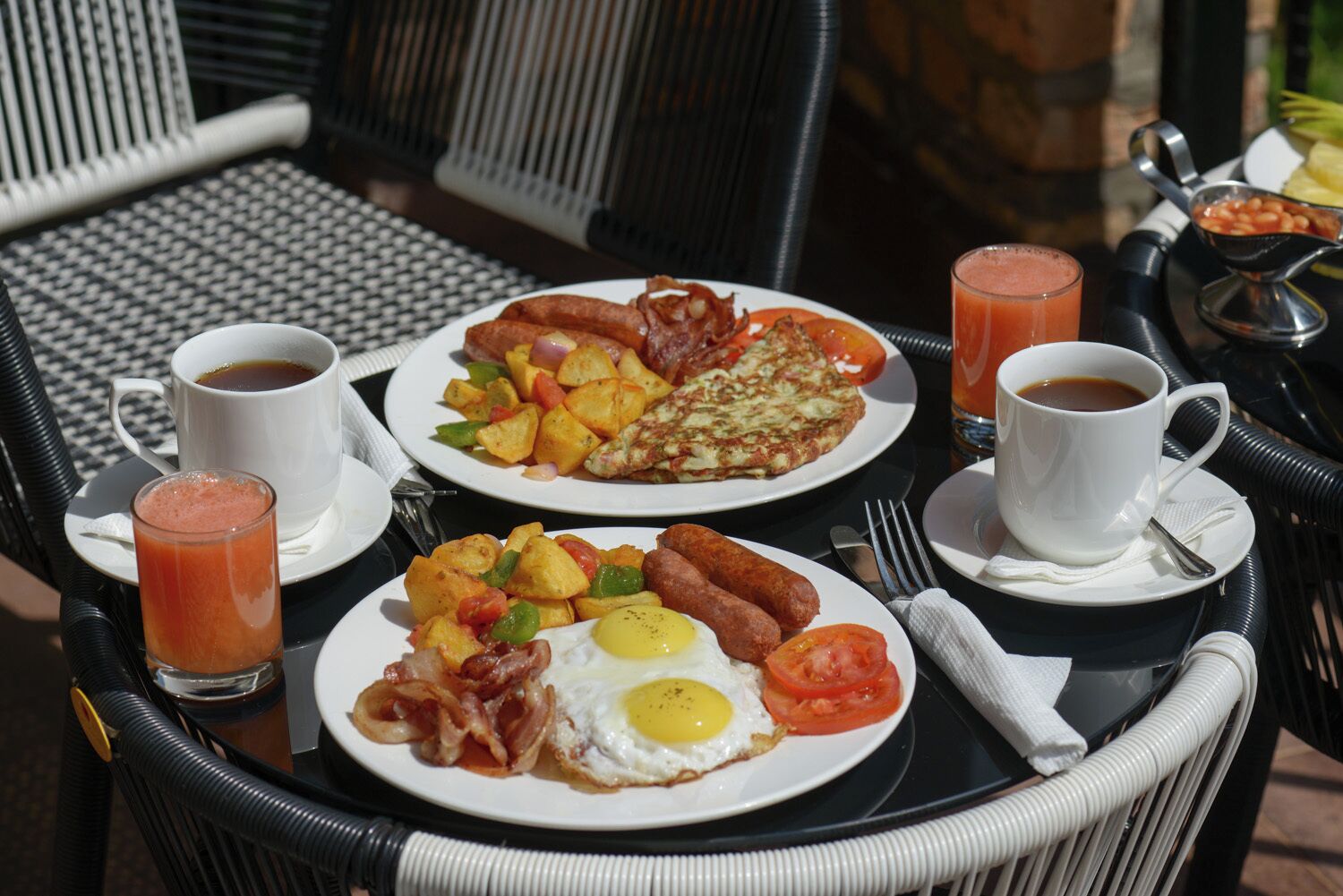 A breakfast setup with plates of eggs, sausages, potatoes, and bacon, accompanied by two cups of coffee and a glass of juice.