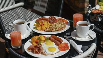 A breakfast setup with plates of eggs, sausages, potatoes, and bacon, accompanied by two cups of coffee and a glass of juice.