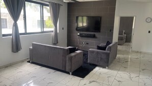 A modern living room with a grey sofa, a smaller armchair, and a flat-screen TV mounted on a brick wall. - Level Suites #3 Premium Suite (Sosúa)