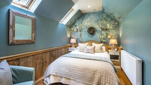 A bedroom with a sloped ceiling, a bed with a quilted bedspread, two bedside tables with lamps, a nightstand, a mirror on the wall, and a window with a view of trees. - The Birdhouse - a luxurious cottage with log burner in the Highlands (Applecross Strathcarron)