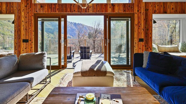 Amplia sala de estar con un gran ventanal corredero que da a una terraza, paneles de madera, un sofá, una mesa de centro y vistas a montañas.
