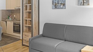 Un salon moderne avec un canapé gris, un plancher en bois et un coin cuisine visible à travers une porte ouverte.