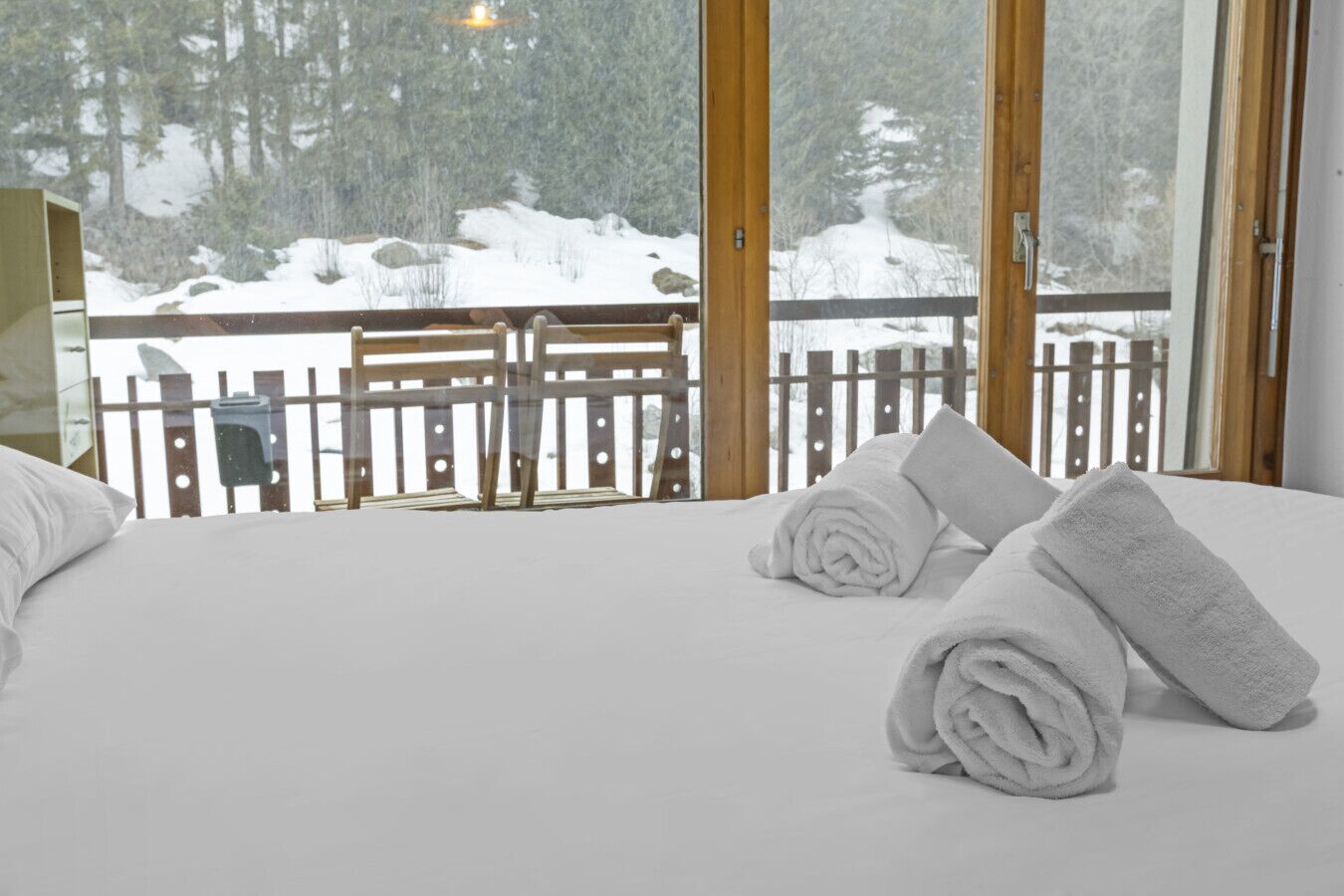 A hotel room with a bed, two rolled towels, a wooden railing, and a snowy view outside.