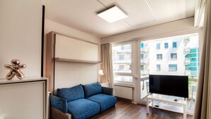 A modern hotel room with a sofa, TV, and large windows offering a view of the city.
