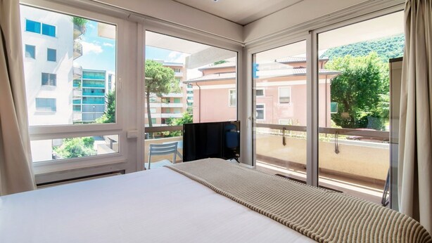 A hotel room with a bed, a TV, and large windows offering a view of the surrounding buildings and greenery.