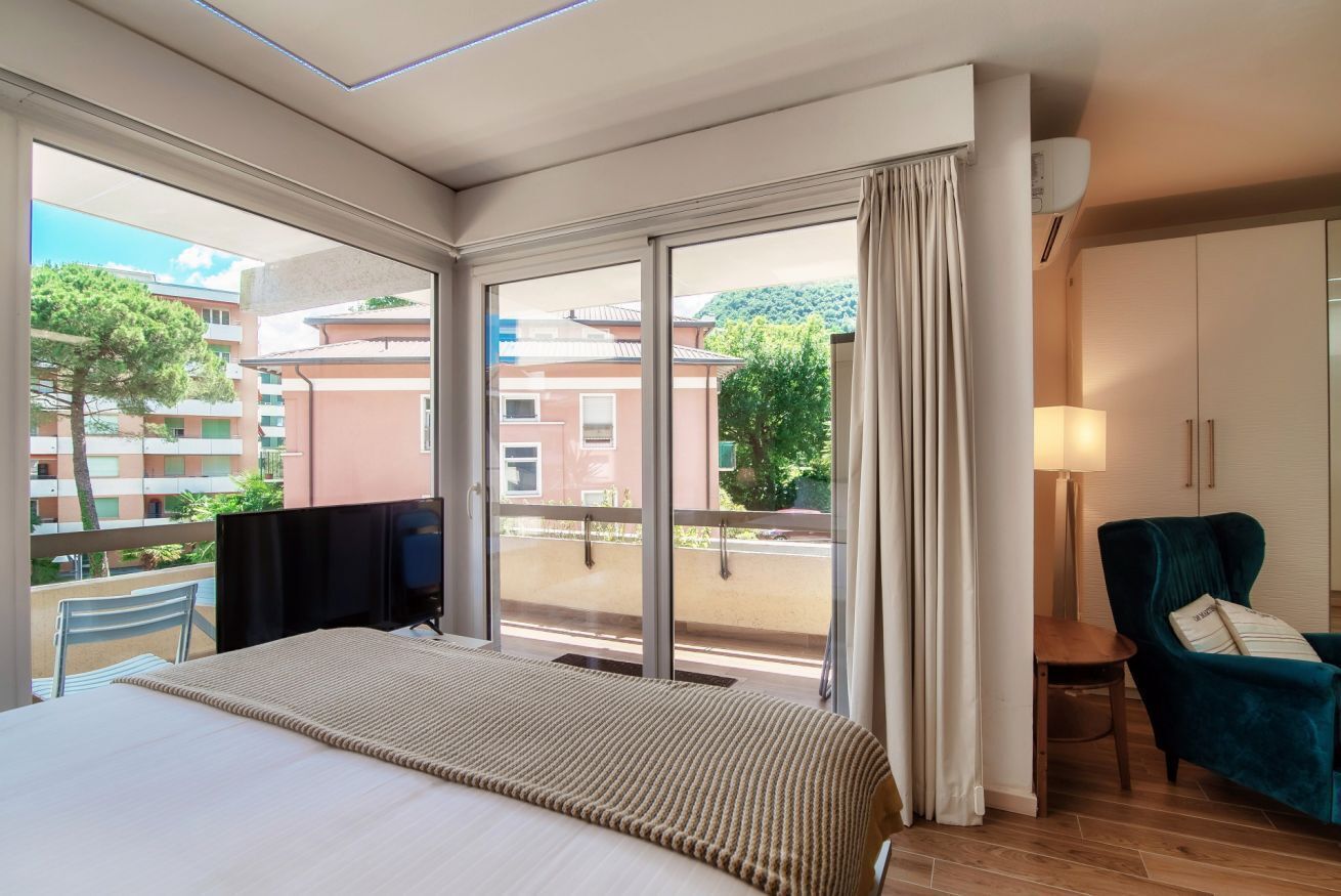 Une chambre d’hôtel avec un lit, une chaise, une télévision et un balcon donnant sur des immeubles et des arbres.