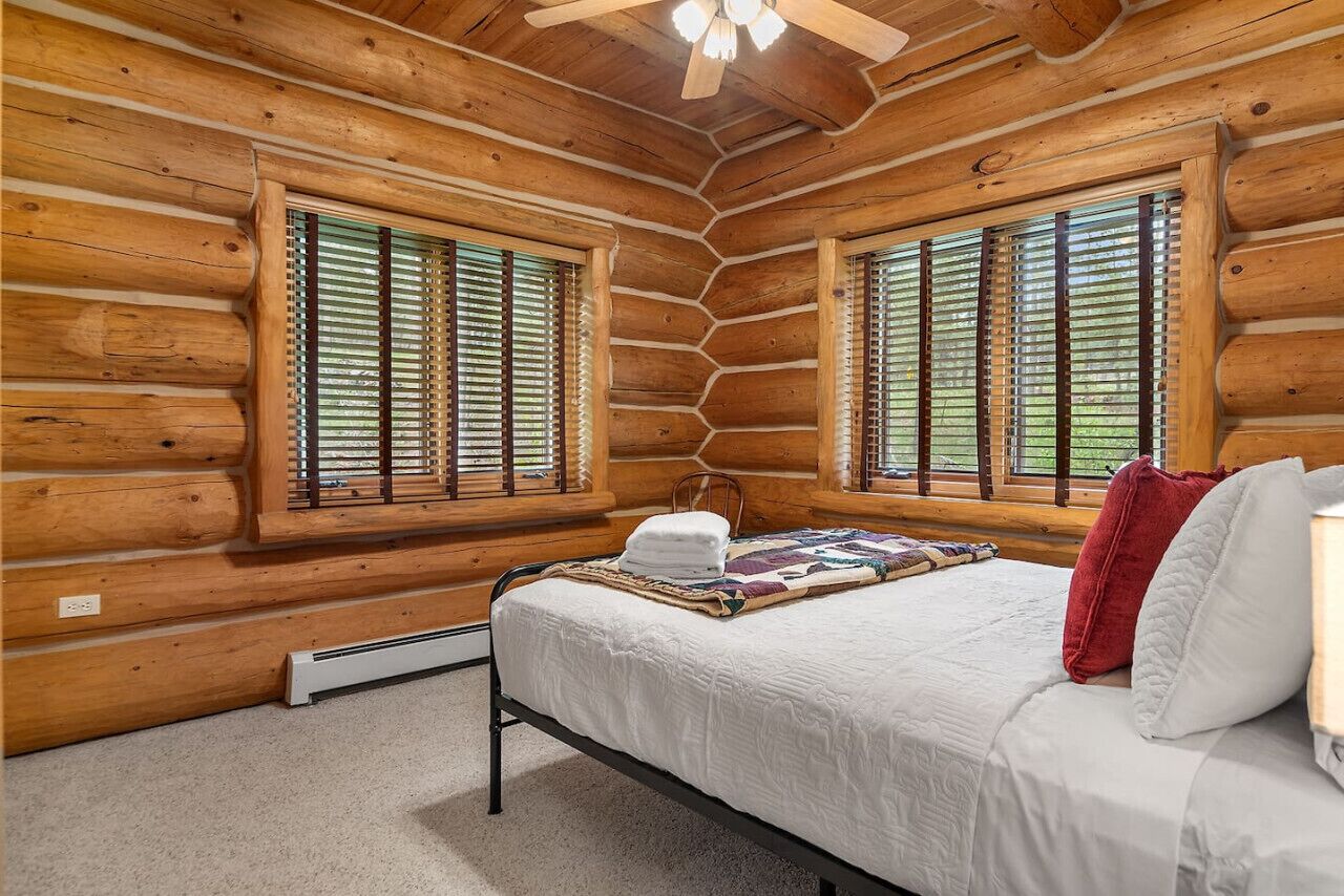 A log cabin bedroom with a bed, two windows with blinds, a ceiling fan, and a heater.