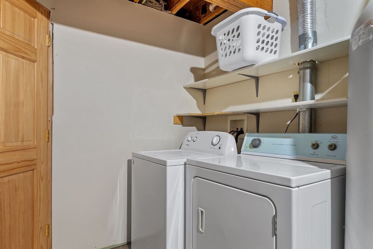 A laundry room with two white front-loading washers and dryers, a white laundry basket on a shelf, and a wooden door.
