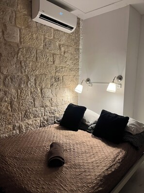 Ein Zimmer mit Steinwand, einem Bett mit zusammengerollter Decke, zwei Wandleuchten und einer Klimaanlage.