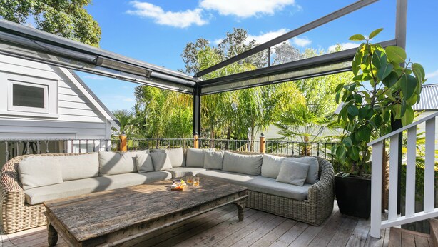 A covered outdoor seating area with a wooden table, wicker furniture, and potted plants.