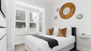 A bedroom with a bed, white walls, a window with blinds, and decorative wall mirrors.