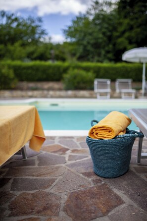 Eine Steinterrasse mit Tisch und Stühlen, ein gewebter Korb mit einem Handtuch und ein Pool im Hintergrund.