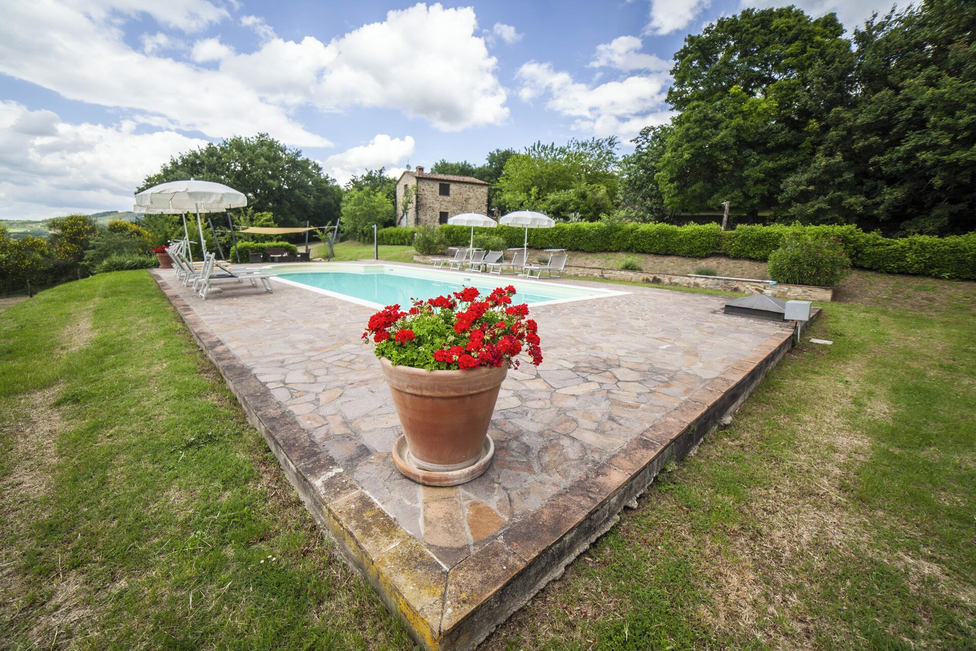 A stone patio with a swimming pool, surrounded by greenery and a stone building.