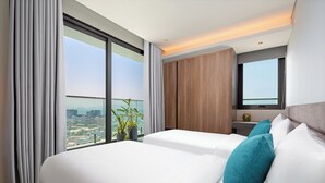 Habitación de hotel con dos camas, un ventanal grande con vistas a la ciudad, un armario de madera y una mesita con un florero.
