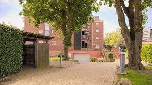 A paved pathway leading to a gated entrance with a small shelter, surrounded by trees and a brick building with balconies.