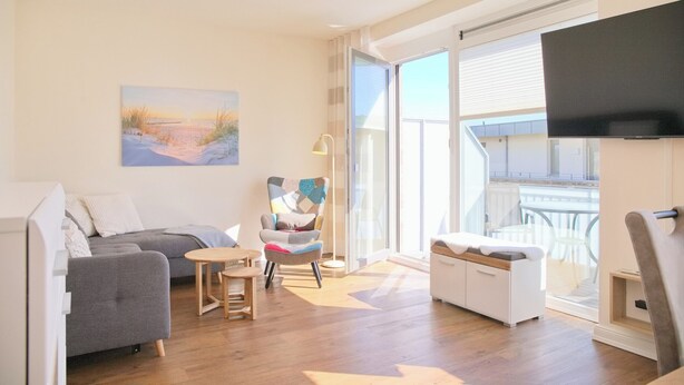 A modern living room with a grey sofa, a small table, a colorful chair, and a white ottoman. There is a large window with a view of the outside.