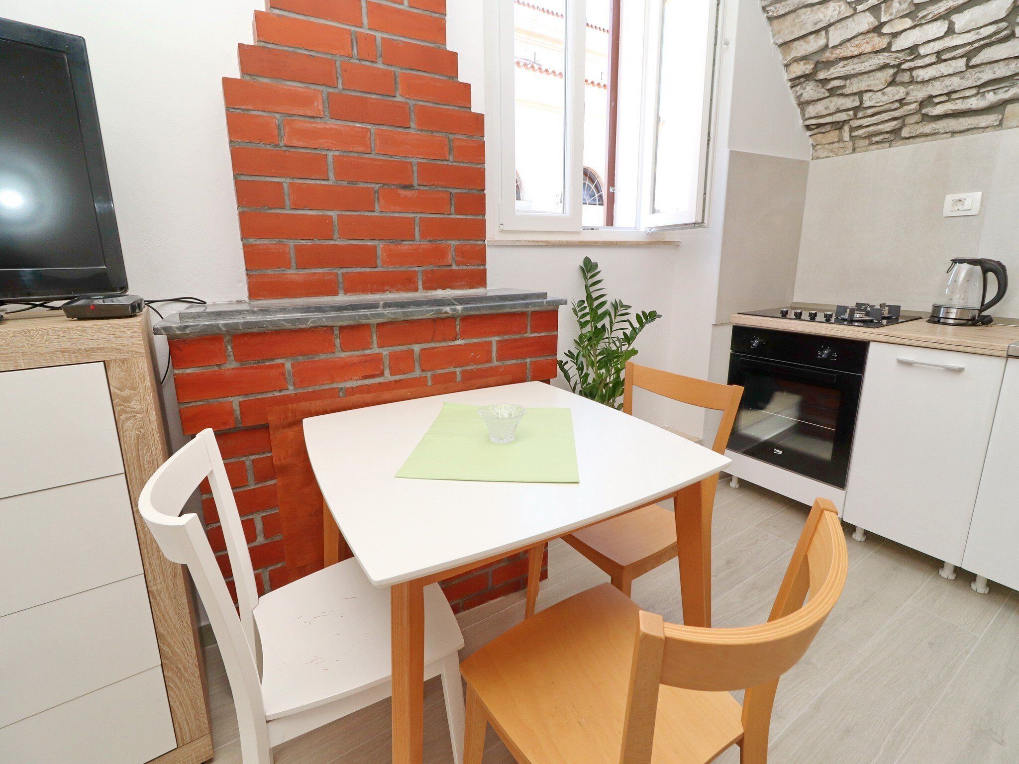 Uma cozinha compacta com parede de tijolos, uma mesa de jantar pequena com cadeiras, uma televisão e um fogão.