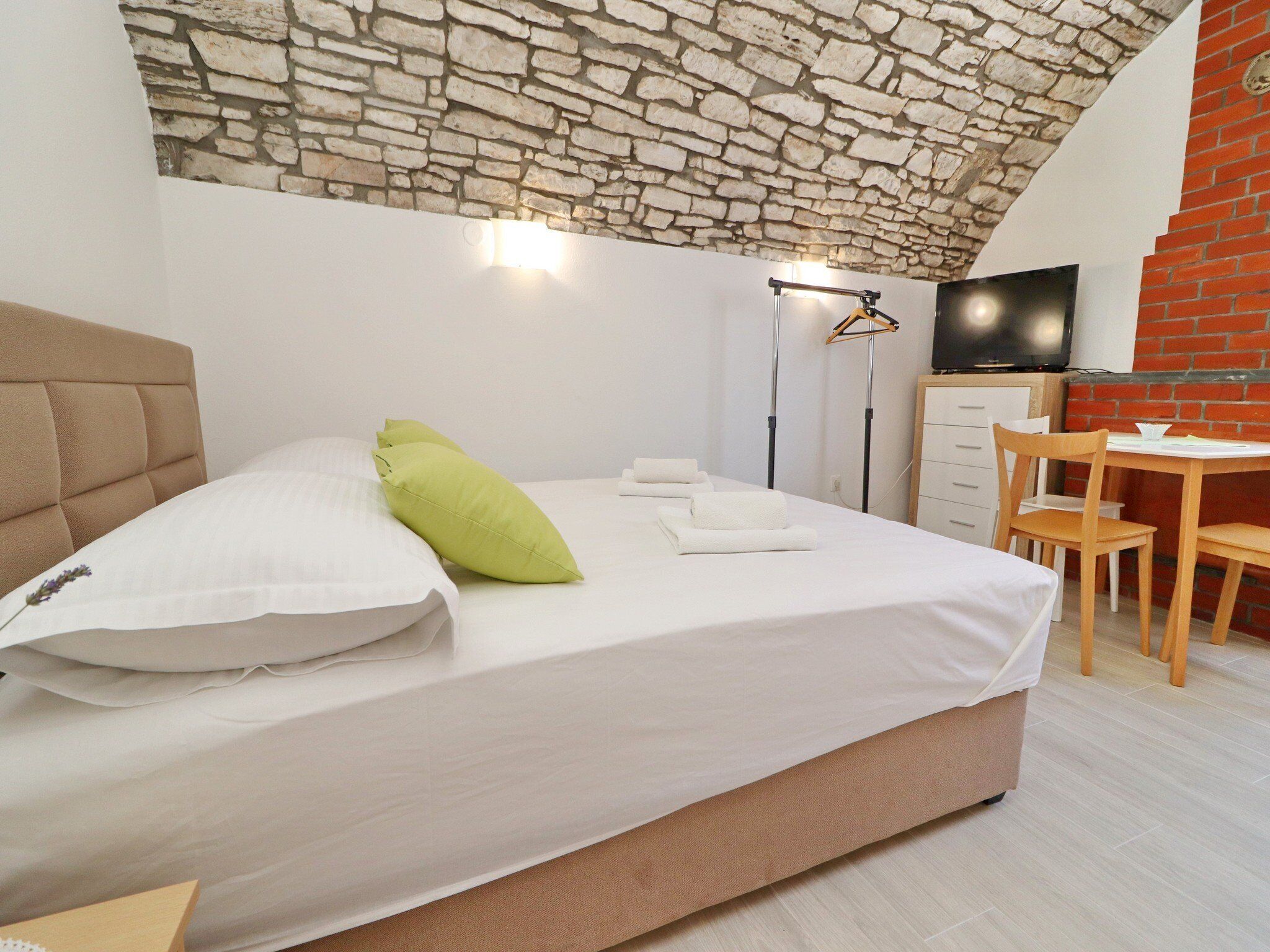 Un dormitorio con techo de piedra, una cama con ropa de cama blanca y almohada verde, una silla de madera, una mesita y un televisor en una pared de ladrillo.