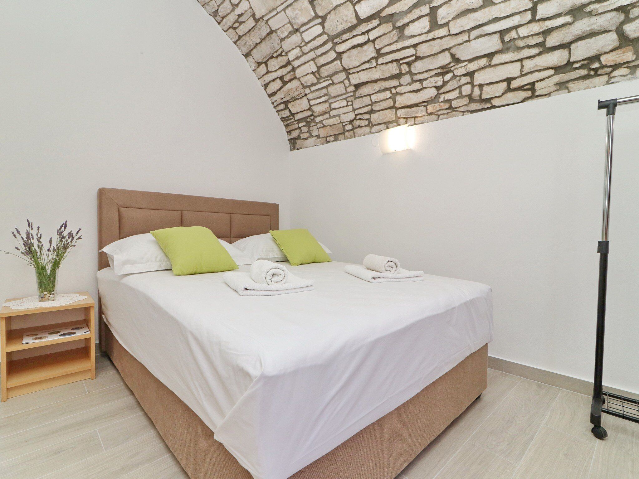 A neatly made bed with white and green pillows, a bedside table with a vase of flowers, and a stone wall above the bed.
