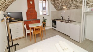 A compact kitchen with a stone ceiling, a brick fireplace, a dining area with a table and chairs, and a small TV on a stand. - Studio Joković (Vela Luka)