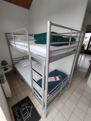 A bunk bed with white bedding and a green blanket, a small table with a lamp, and a doormat with a house symbol.