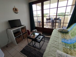 A room with a bed, a TV on a stand, a small table, a window with a view of the outside, and a balcony with chairs. - 5 min from the ocean / free shuttle service Furnished studio in a family residence (Talmont-Saint-Hilaire)