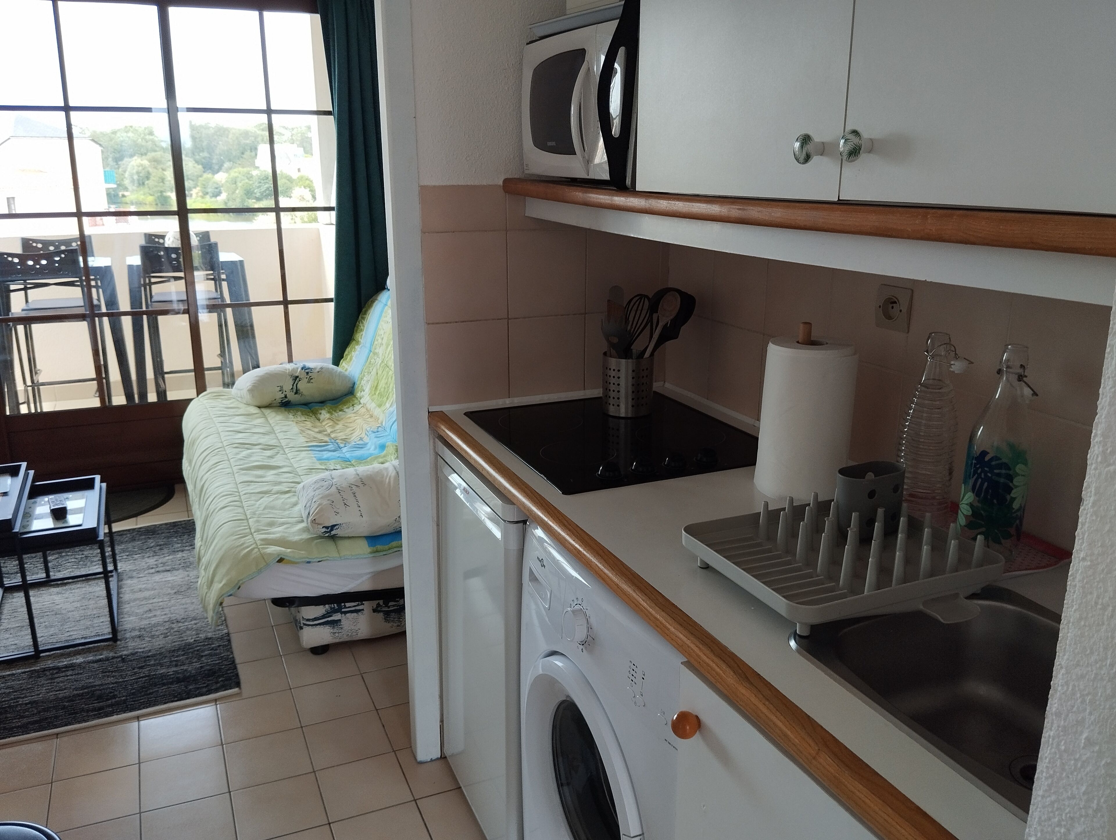 Ein kleines Hotelzimmer mit Bett, Waschmaschine, Mikrowelle, Spüle und einem Fenster mit Blick auf Grünflächen.