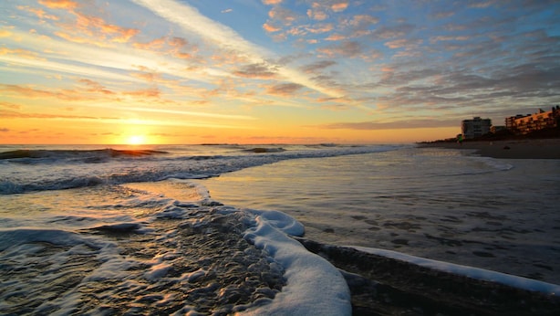 Une plage avec des vagues au coucher du soleil, des bâtiments en arrière-plan et un ciel dégagé.
